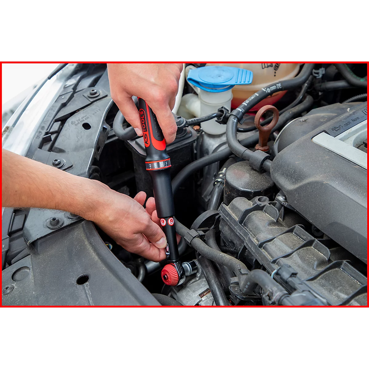 Person verschraubt mit rotem und schwarzem Werkzeug im Motorraum eines Autos.